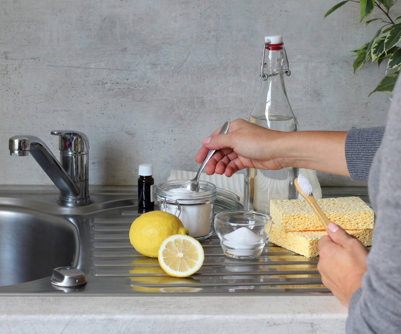 Hand holding spoon with white power next to jar of clear lquid with lemons and cleaning cloths in background