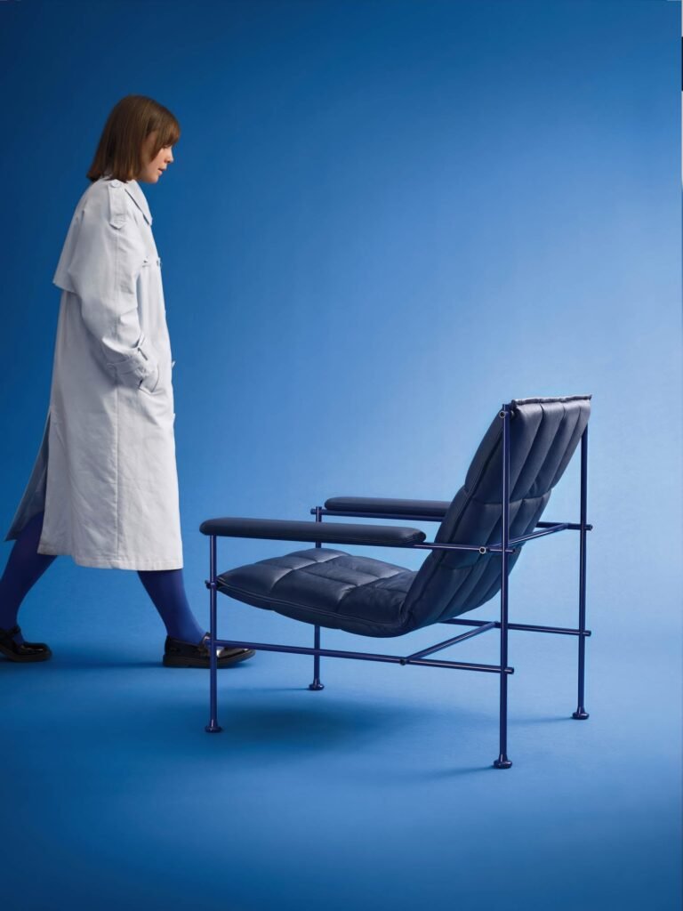A woman in a light-colored coat walks past a modern, blue cushioned chair against a plain blue background.
