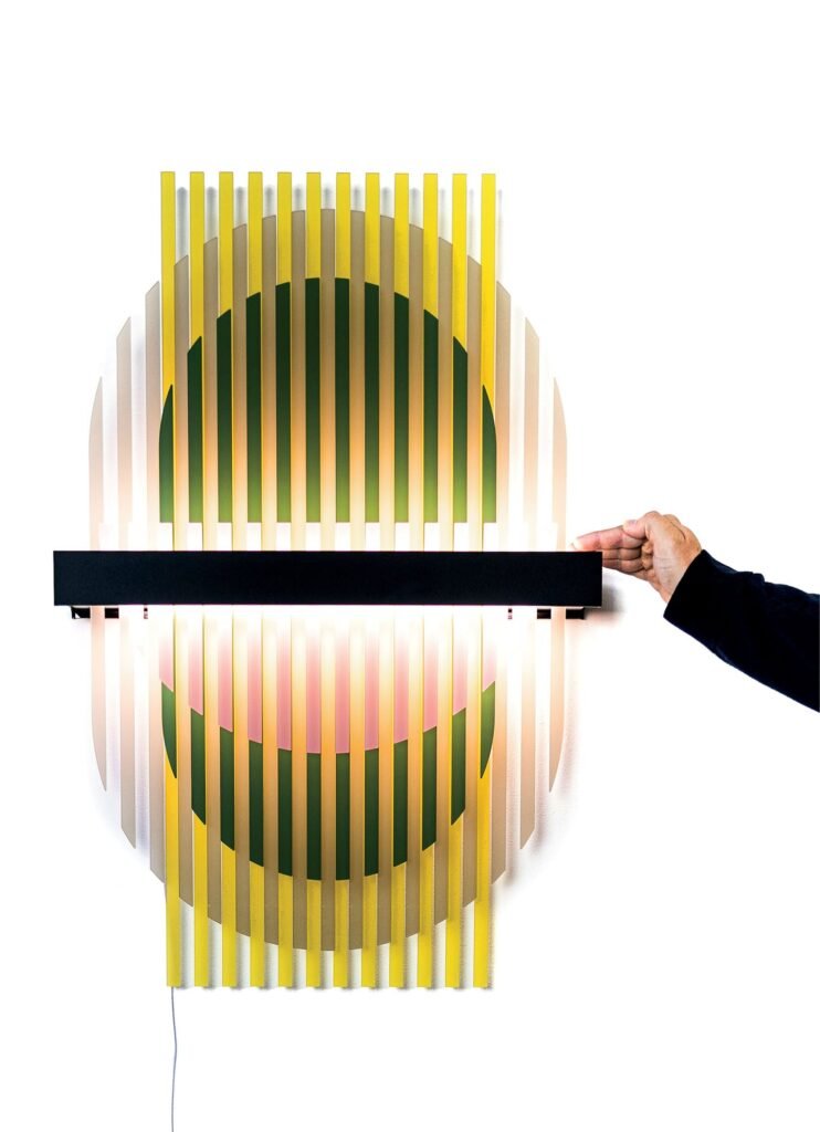 A hand adjusts a horizontal black bar on a circular wall-mounted light fixture with vertical yellow and white slats and a glowing center.