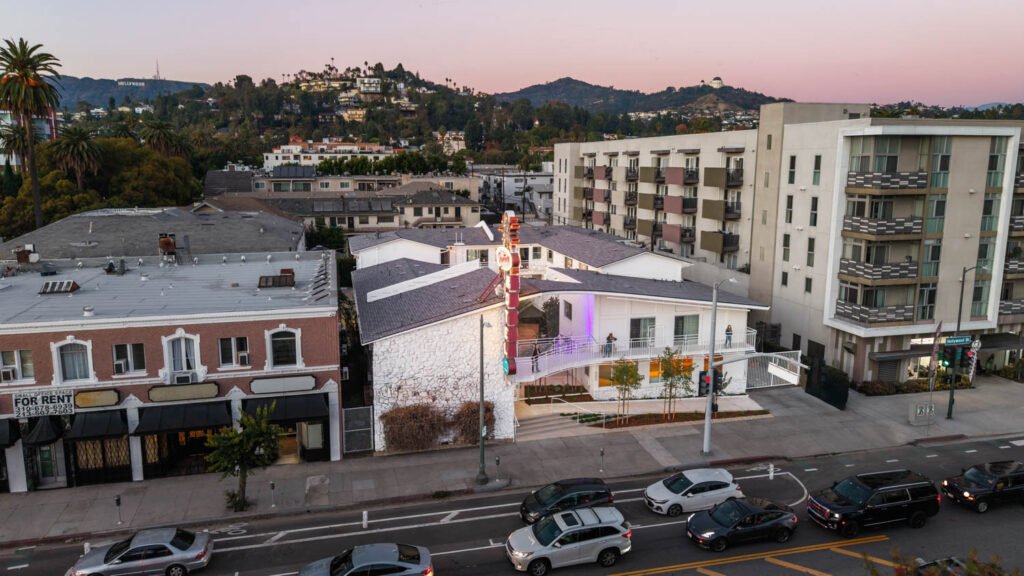 Aerial view of a mid-century motel on a busy street, surrounded by apartments and hills in the background at dusk.