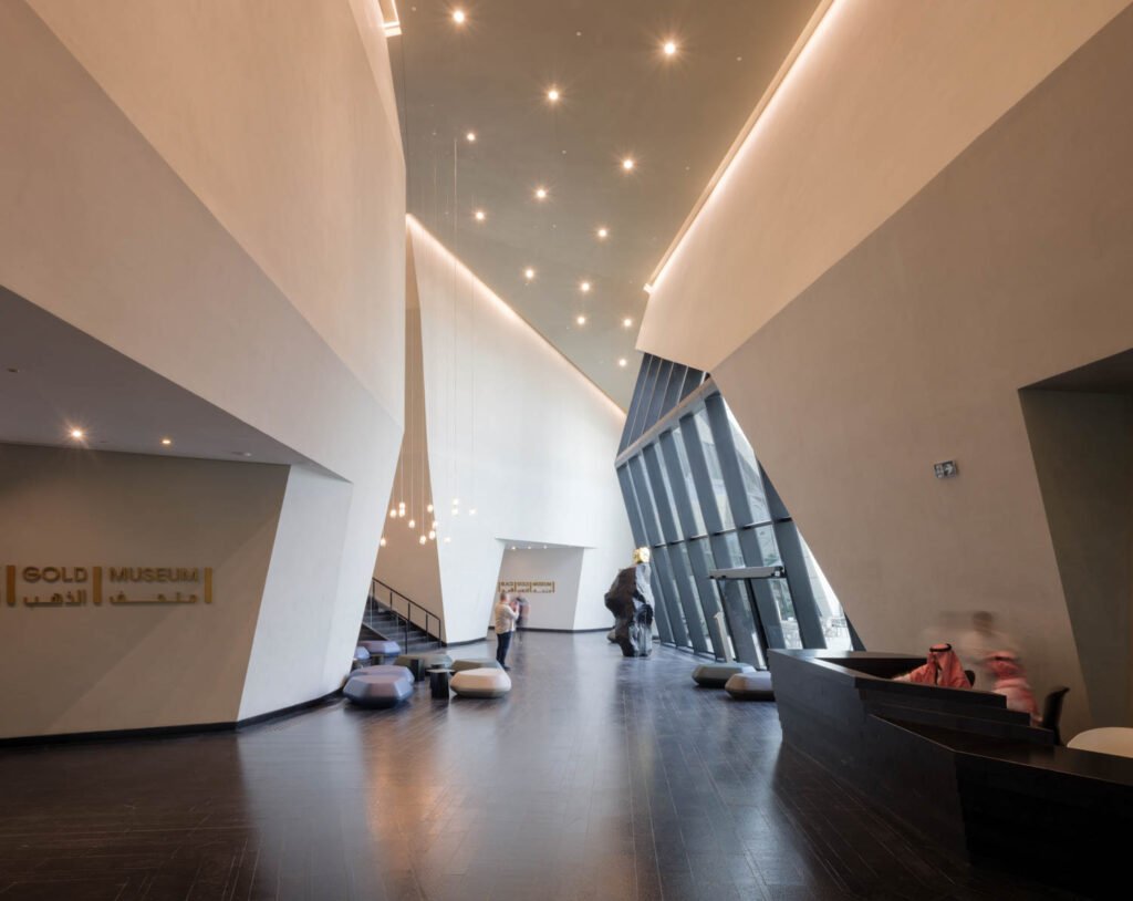 Spacious museum lobby with modern angular white walls, dark wood floor, scattered seating, and visitors walking and sitting under ceiling lights.