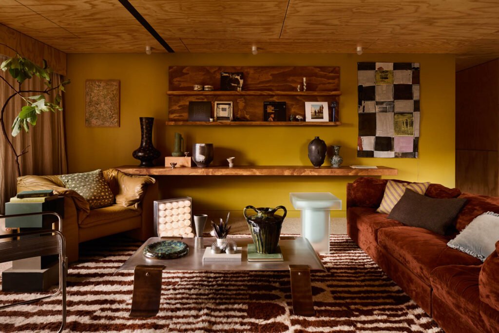 Living room with mustard yellow walls, wood shelves, eclectic decor, brown and yellow sofas, patterned rug, and a modern coffee table with vases and books.