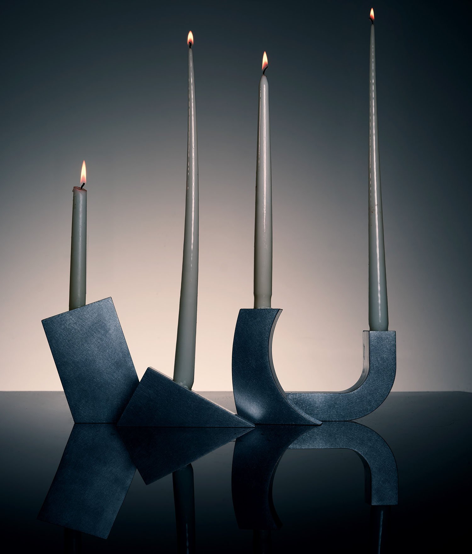 Three modern, geometric metal candle holders with tall lit taper candles are arranged on a reflective surface against a gradient background.